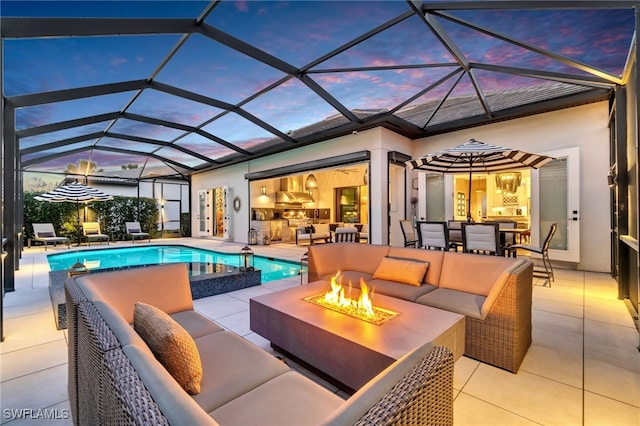 pool at dusk with an outdoor kitchen, glass enclosure, a patio area, and an outdoor living space with a fire pit