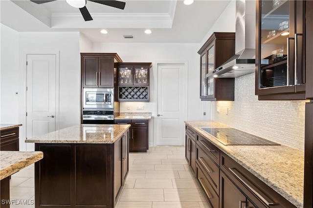 kitchen with a center island, appliances with stainless steel finishes, a tray ceiling, light stone countertops, and wall chimney range hood