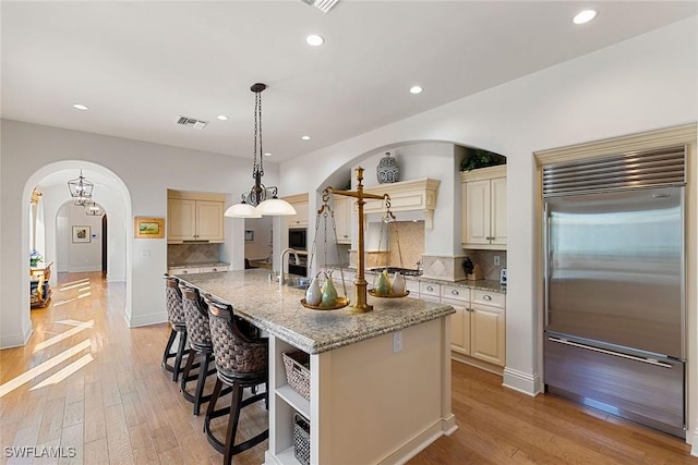 kitchen featuring built in appliances, arched walkways, light stone counters, cream cabinets, and a kitchen island with sink