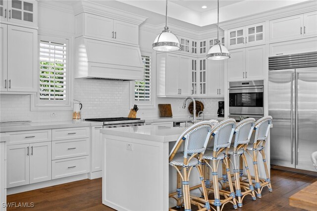 kitchen with stainless steel appliances, custom exhaust hood, a center island with sink, and white cabinetry