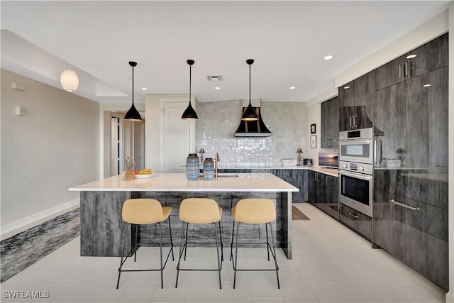 kitchen with wall chimney range hood, hanging light fixtures, backsplash, a center island with sink, and stainless steel double oven