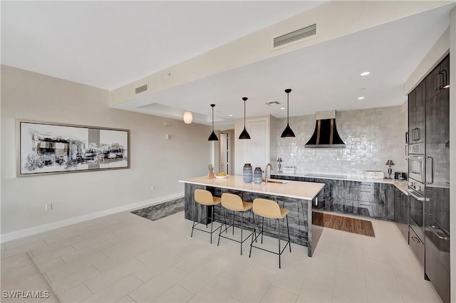 kitchen featuring wall chimney exhaust hood, a kitchen bar, stainless steel oven, pendant lighting, and a kitchen island with sink