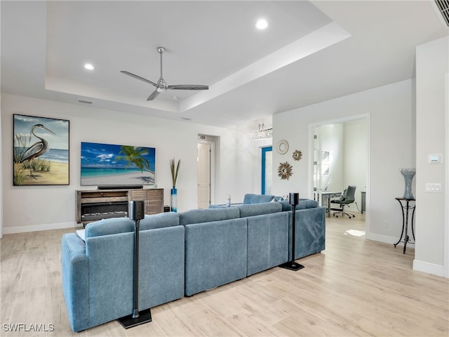 living room with a raised ceiling, ceiling fan, and light wood-type flooring