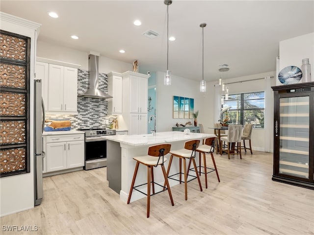 kitchen featuring white cabinetry, an island with sink, appliances with stainless steel finishes, and wall chimney range hood