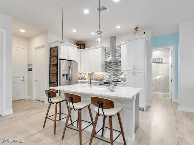 kitchen featuring pendant lighting, stainless steel appliances, white cabinets, a center island with sink, and wall chimney exhaust hood