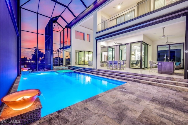 pool at dusk featuring glass enclosure, a patio area, pool water feature, and ceiling fan