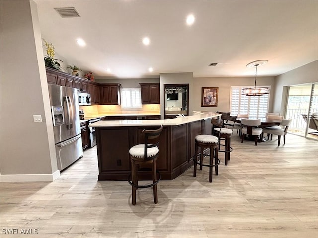 kitchen featuring appliances with stainless steel finishes, an island with sink, hanging light fixtures, dark brown cabinetry, and light hardwood / wood-style flooring