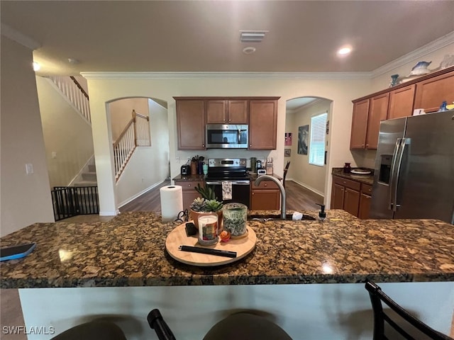 kitchen with crown molding, dark wood-type flooring, appliances with stainless steel finishes, dark stone countertops, and a kitchen bar