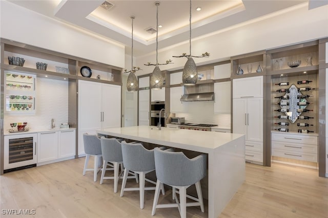 kitchen featuring white cabinetry, hanging light fixtures, wine cooler, a raised ceiling, and a large island with sink