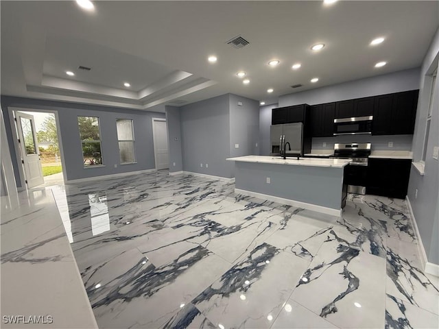 kitchen with sink, appliances with stainless steel finishes, light stone counters, a center island with sink, and a raised ceiling