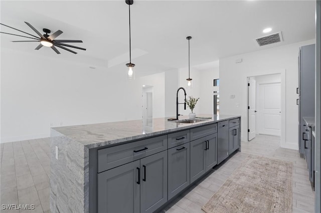 kitchen featuring sink, gray cabinetry, decorative light fixtures, an island with sink, and light stone countertops