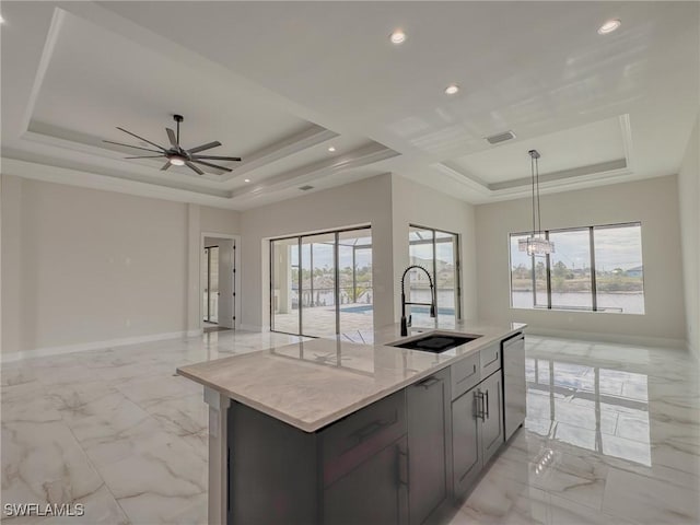 kitchen with sink, a center island with sink, a raised ceiling, pendant lighting, and light stone countertops