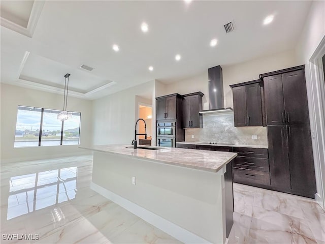 kitchen featuring sink, wall chimney exhaust hood, an island with sink, a raised ceiling, and stainless steel double oven