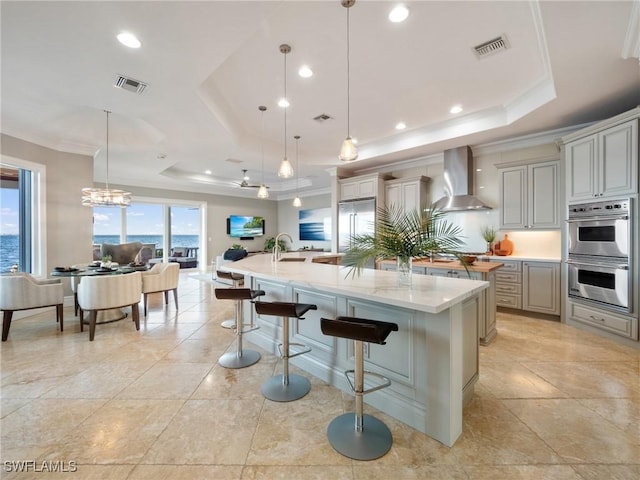 kitchen featuring a large island with sink, a tray ceiling, a breakfast bar area, and wall chimney exhaust hood