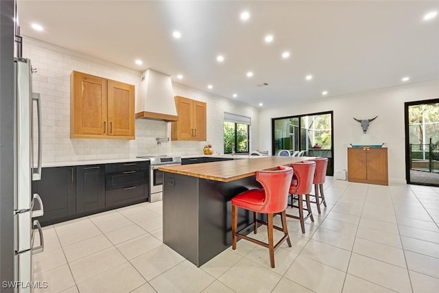 kitchen with butcher block countertops, custom range hood, a center island, appliances with stainless steel finishes, and light tile patterned flooring