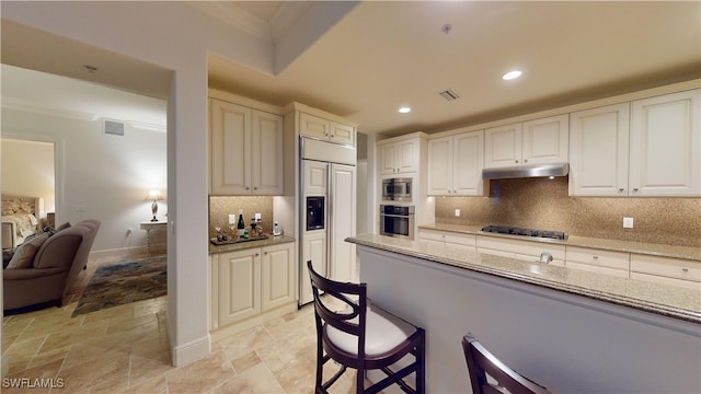 kitchen featuring a breakfast bar, backsplash, built in appliances, ornamental molding, and light stone countertops