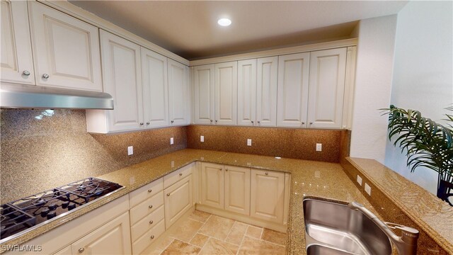 kitchen with black gas stovetop, under cabinet range hood, stone countertops, a sink, and decorative backsplash