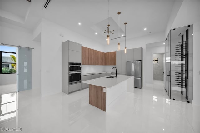 kitchen with appliances with stainless steel finishes, a towering ceiling, sink, hanging light fixtures, and a center island with sink