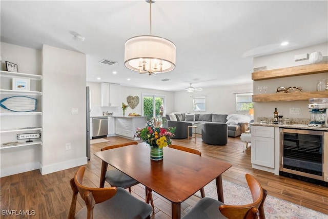 dining room featuring indoor bar, beverage cooler, ceiling fan with notable chandelier, and light wood-type flooring