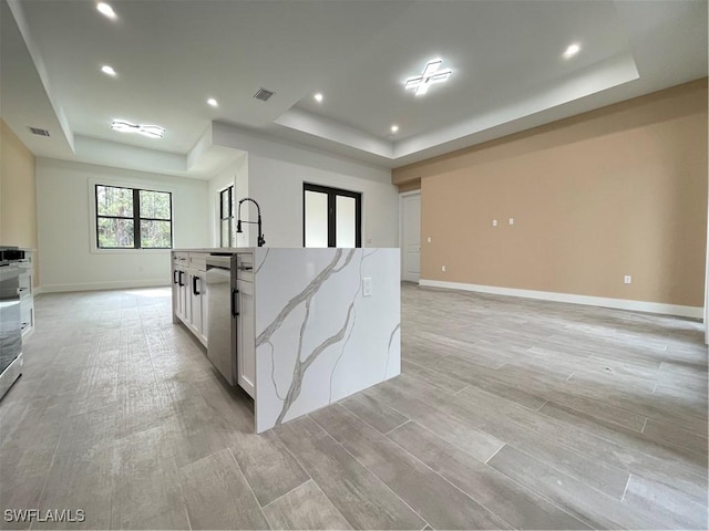 kitchen featuring white cabinetry, light stone countertops, a center island with sink, stainless steel dishwasher, and a raised ceiling