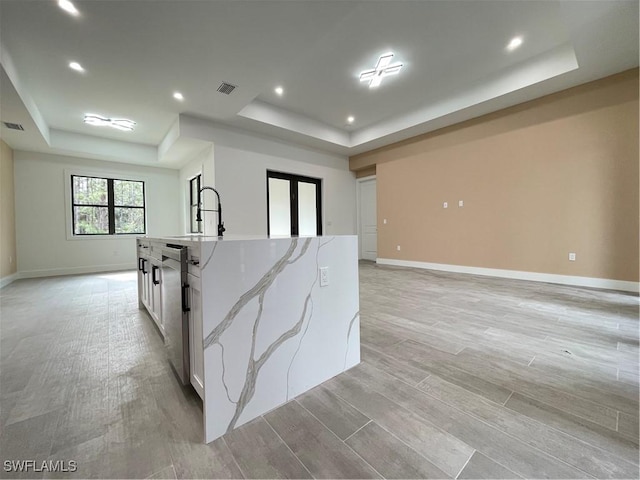 kitchen featuring white cabinetry, light hardwood / wood-style flooring, a raised ceiling, light stone countertops, and a kitchen island with sink