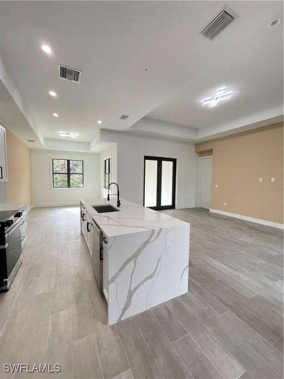 kitchen featuring sink, light stone countertops, white cabinets, a center island with sink, and a raised ceiling