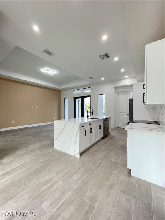 kitchen with sink, white cabinets, a tray ceiling, light stone countertops, and a center island with sink