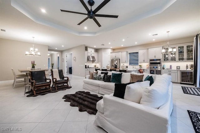 tiled living room with bar, beverage cooler, a tray ceiling, and ceiling fan with notable chandelier