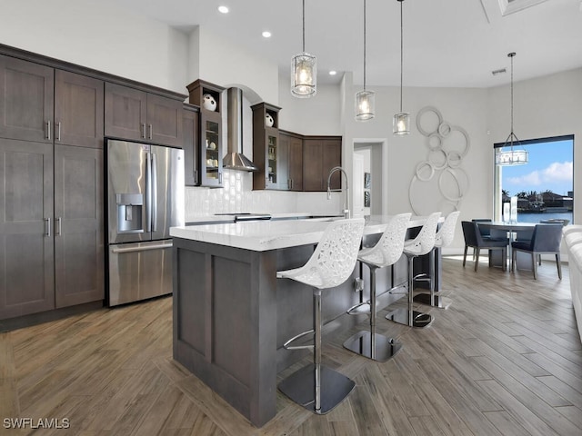 kitchen featuring a kitchen island with sink, dark brown cabinetry, a kitchen bar, stainless steel fridge with ice dispenser, and wall chimney exhaust hood