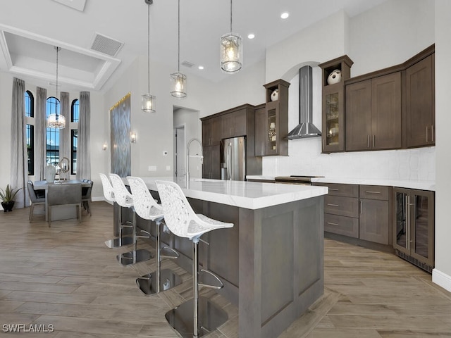 kitchen with decorative light fixtures, appliances with stainless steel finishes, an island with sink, beverage cooler, and wall chimney range hood