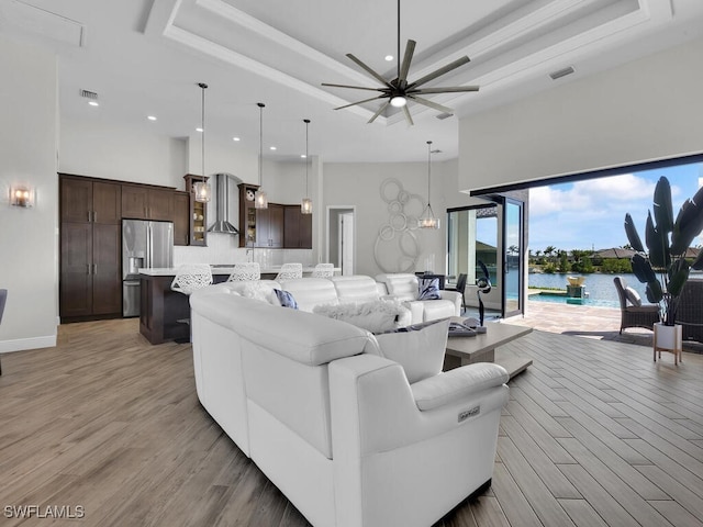 living room featuring light hardwood / wood-style flooring, a towering ceiling, a water view, ceiling fan with notable chandelier, and a raised ceiling