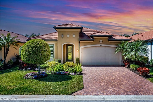 mediterranean / spanish house featuring decorative driveway, a yard, stucco siding, an attached garage, and a tiled roof