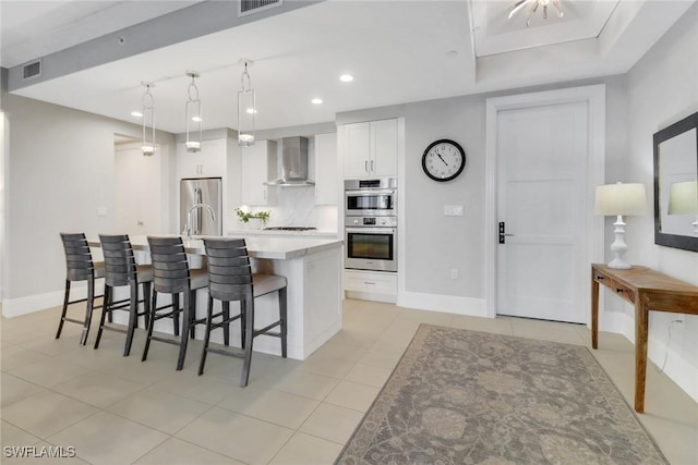 kitchen featuring appliances with stainless steel finishes, white cabinetry, hanging light fixtures, a center island with sink, and wall chimney exhaust hood