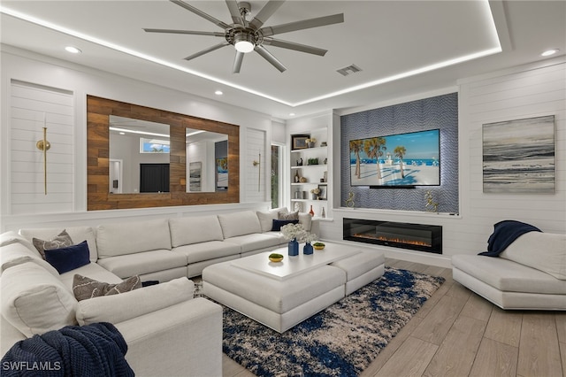 living room with ceiling fan, wooden walls, a tray ceiling, light hardwood / wood-style floors, and built in shelves