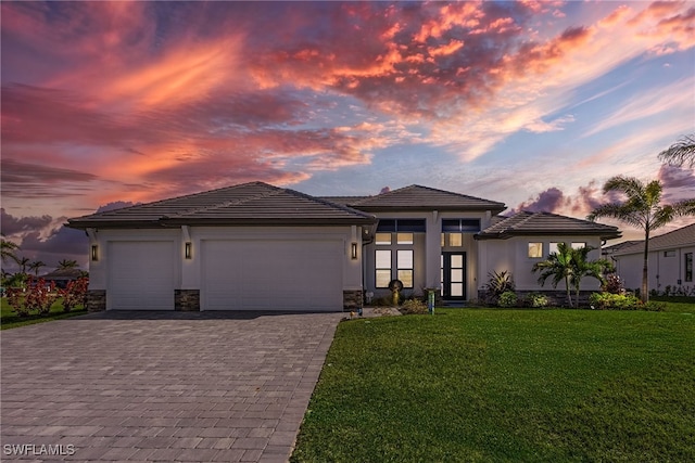 prairie-style home with a lawn, stone siding, an attached garage, decorative driveway, and stucco siding