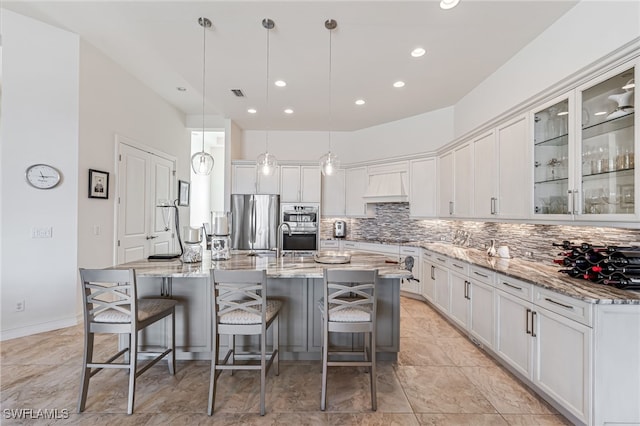 kitchen featuring an island with sink, appliances with stainless steel finishes, premium range hood, and decorative light fixtures