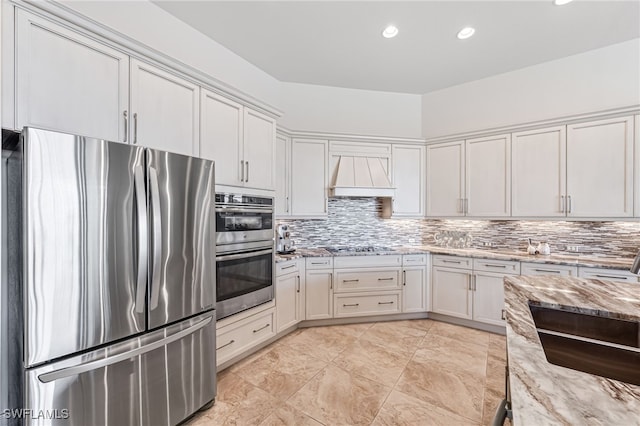 kitchen featuring light stone counters, appliances with stainless steel finishes, custom range hood, white cabinets, and backsplash