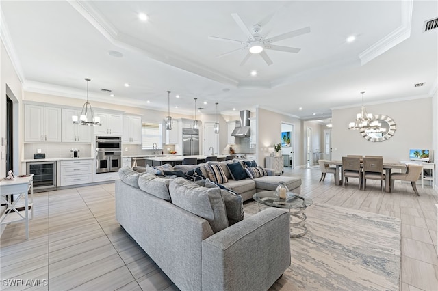 living room with ceiling fan with notable chandelier, sink, beverage cooler, ornamental molding, and a raised ceiling