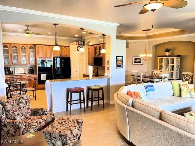 living room with crown molding, ceiling fan with notable chandelier, a tray ceiling, and light tile patterned flooring