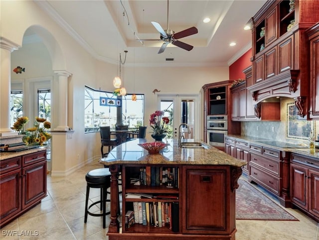 kitchen featuring sink, a tray ceiling, an island with sink, black electric cooktop, and ornate columns