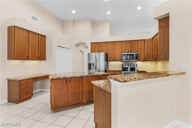 kitchen with light tile patterned floors, appliances with stainless steel finishes, light stone countertops, an island with sink, and kitchen peninsula