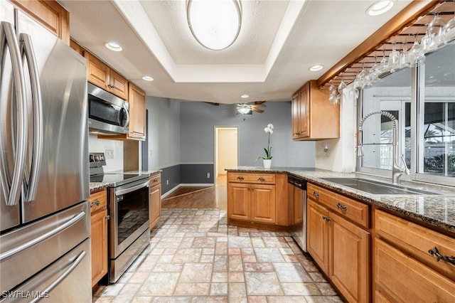 kitchen with sink, stainless steel appliances, a tray ceiling, kitchen peninsula, and dark stone counters