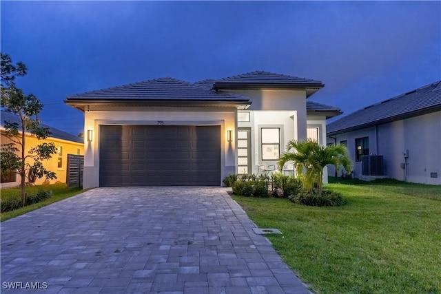 prairie-style home with an attached garage, decorative driveway, a front lawn, central AC, and stucco siding