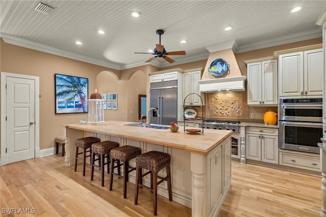 kitchen with butcher block counters, tasteful backsplash, an island with sink, stainless steel appliances, and cream cabinets