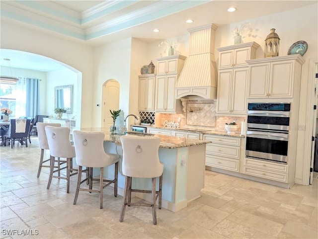 kitchen with arched walkways, light stone counters, stone tile floors, cream cabinetry, and custom range hood