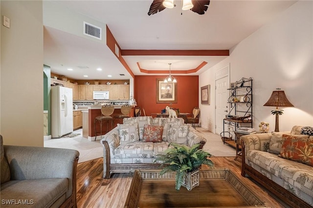 living area with ceiling fan with notable chandelier, a tray ceiling, visible vents, and light wood-style floors