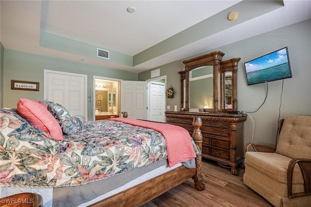 bedroom featuring ensuite bath, visible vents, a raised ceiling, and wood finished floors