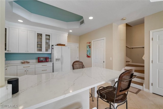 kitchen with a kitchen bar, white cabinetry, light stone counters, a tray ceiling, and white refrigerator with ice dispenser