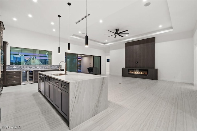 kitchen featuring sink, a kitchen island with sink, hanging light fixtures, light stone countertops, and a raised ceiling