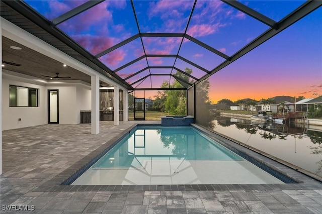 pool at dusk with a jacuzzi, ceiling fan, glass enclosure, a water view, and a patio area